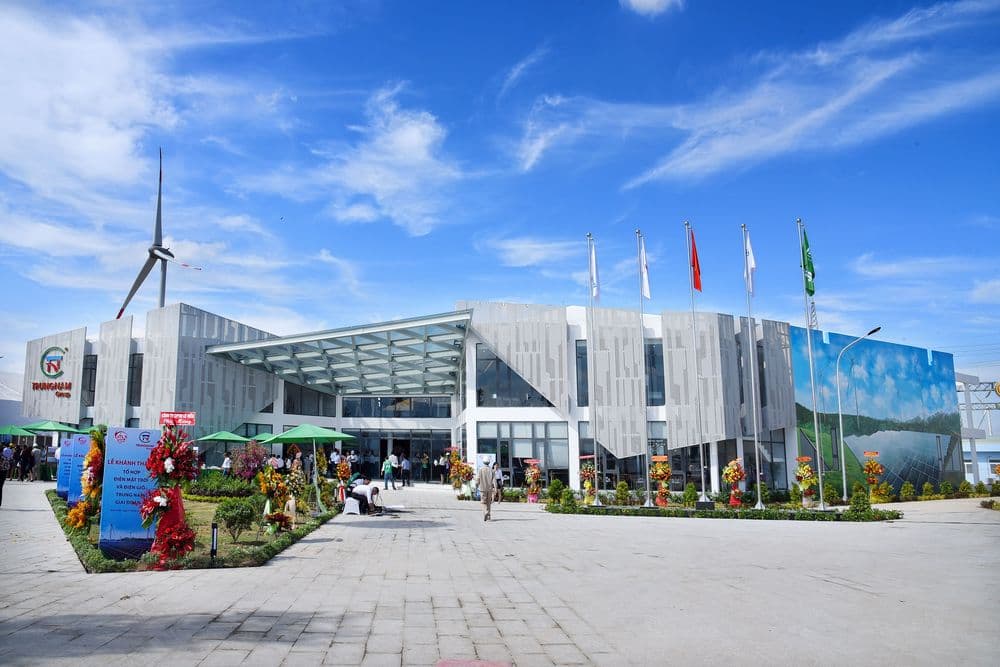 Modern exhibition building with glass facade, flags, and a wind turbine under a blue sky.