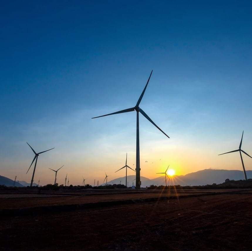 Wind turbines silhouetted against a vibrant sunset sky over a dark field.