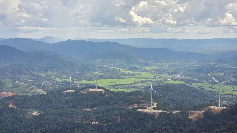 Wind turbines rise from forested mountain peaks, generating renewable energy in a lush green valley.